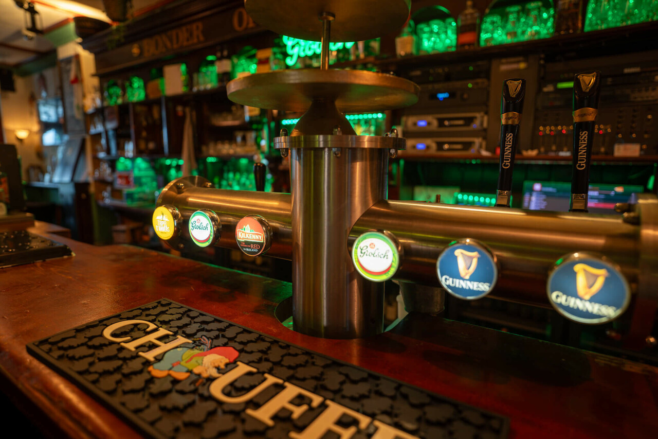 _dsc1915-enhanced-nr.jpg A bar counter features taps with various beer brands, including Heineken, Kilkenny, and Guinness. The backdrop is illuminated with green lighting, showcasing shelves filled with glassware. A black mat with colorful lettering is displayed on the counter.