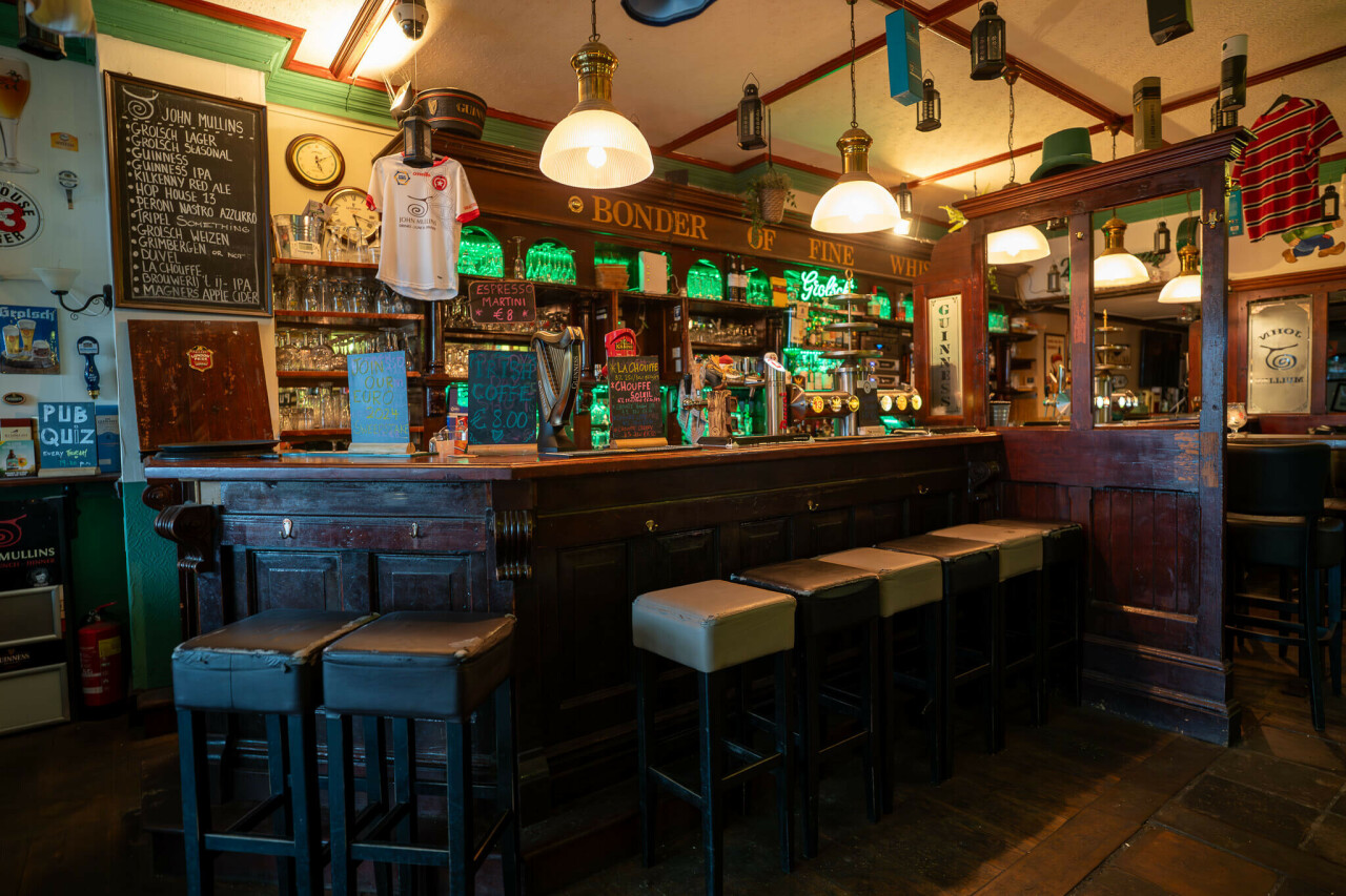 _dsc1934-enhanced-nr.jpg A cozy pub interior features a long wooden bar with high-backed stools, illuminated by pendant lights. Shelves behind the bar display green bottles, and chalkboards showcase drink specials. A football jersey hangs decoratively, adding to the inviting atmosphere.