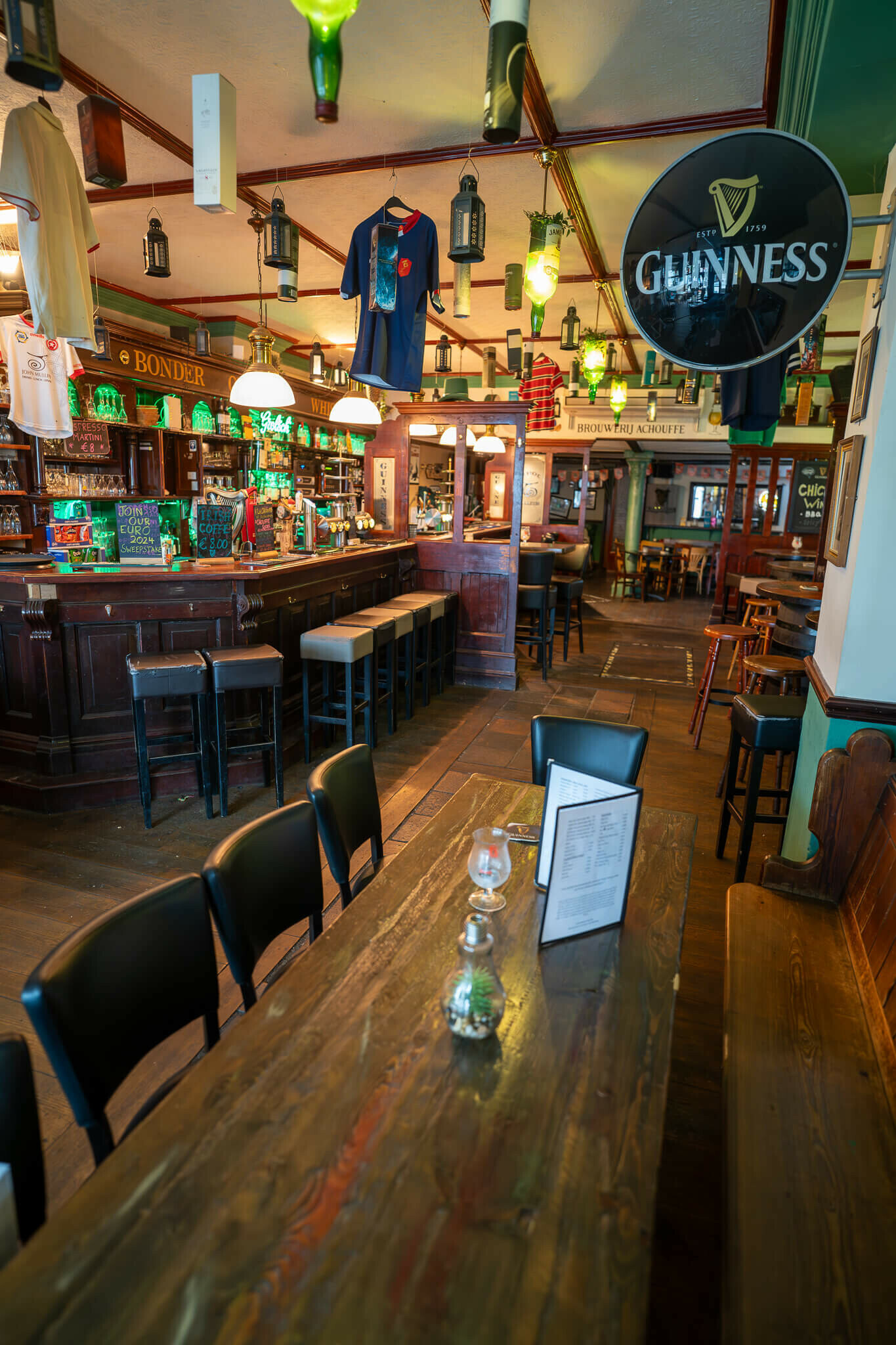 _dsc1950-enhanced-nr.jpg A cozy pub interior features dark wooden furniture and a bar lined with green-lit decorations. Various shirts and bottles hang from the ceiling, while menus and small glassware rest on a long wooden table. The atmosphere is inviting and traditional, with a mix of seating arrangements throughout the space.